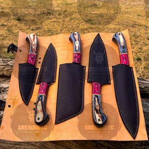 May include: A set of five knives with black blades and multi-colored handles. The handles feature a combination of red, blue, and wood tones. Each knife is displayed with a black leather sheath on a tan surface. The text "SHEROMNYC" is visible.