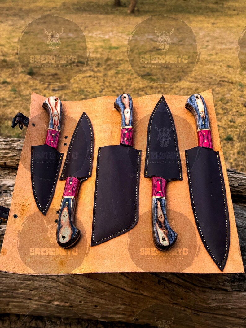 May include: A set of five knives with black blades and multi-colored handles. The handles feature a combination of red, blue, and wood tones. Each knife is displayed with a black leather sheath on a tan surface. The text "SHEROMNYC" is visible.