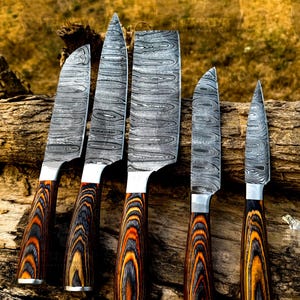 May include: A set of five Damascus steel knives with patterned blades and wooden handles. The handles feature a striking design of black, orange, and brown wood grain. The knives are arranged on a piece of weathered wood.