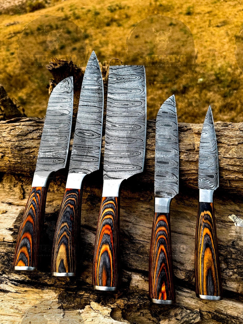 May include: A set of five Damascus steel knives with patterned blades and wooden handles. The handles feature a striking design of black, orange, and brown wood grain. The knives are arranged on a piece of weathered wood.