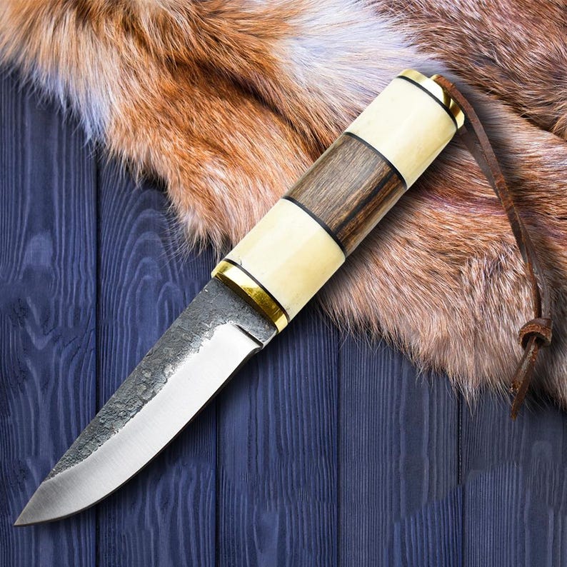 May include: A handcrafted knife with a polished steel blade and a handle featuring alternating sections of light cream, dark wood, and brass accents. A leather strap is attached to the handle. The knife rests on a blue wooden surface.