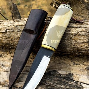 May include: A handcrafted knife with a light-colored handle and a dark, curved blade. The knife rests on a piece of wood next to a dark brown leather sheath. The handle has a gold-colored accent and a leather strap.