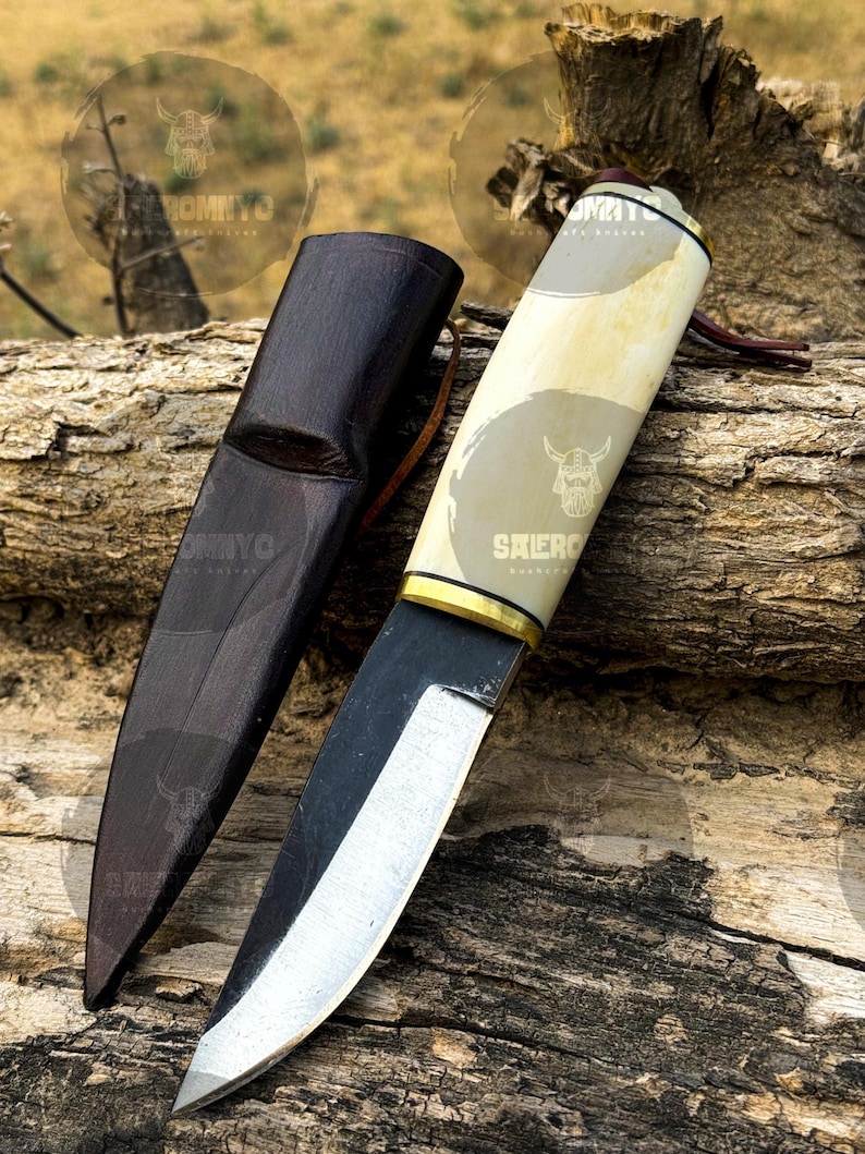 May include: A handcrafted knife with a light-colored handle and a dark, curved blade. The knife rests on a piece of wood next to a dark brown leather sheath. The handle has a gold-colored accent and a leather strap.