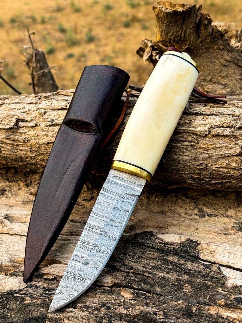 May include: A handcrafted knife with a cream-colored handle and a patterned steel blade, resting beside a dark brown leather sheath. The handle is accented with gold-colored metal rings. The knife is designed for outdoor use.
