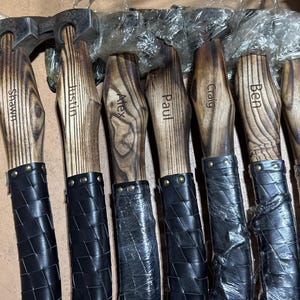 May include: A collection of personalized axes with wooden handles and black leather-wrapped grips. Each axe head is made of metal and the wooden handles are engraved with names: Shawn, Justin, Alex, Paul, Craig, Ben, and Walter.