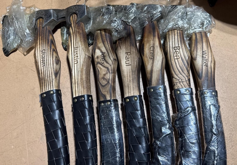 May include: A collection of personalized axes with wooden handles and black leather-wrapped grips. Each axe head is made of metal and the wooden handles are engraved with names: Shawn, Justin, Alex, Paul, Craig, Ben, and Walter.
