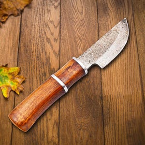 May include: A brown wooden handled knife with a patterned steel blade. The knife is lying on a wooden surface.