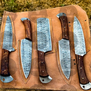 May include: A set of five Damascus steel knives with wooden handles, displayed in a brown leather sheath. The blades have a unique patterned design. The handles are dark brown with a curved design. The sheath is tan with a stitched border.