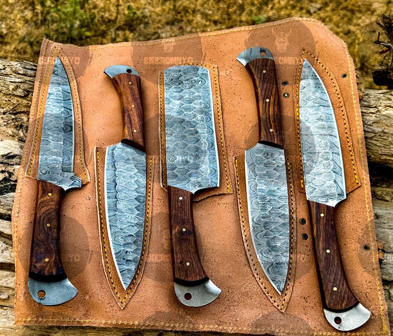 May include: A set of five Damascus steel knives with wooden handles, displayed in a brown leather sheath. The blades have a unique patterned design. The handles are dark brown with a curved design. The sheath is tan with a stitched border.