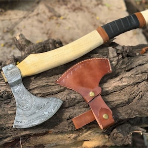 May include: A handcrafted axe with a patterned steel blade and a light wood handle. The handle is wrapped in black and brown leather. A brown leather sheath with brass rivets protects the blade. The axe is displayed on a weathered wooden surface.