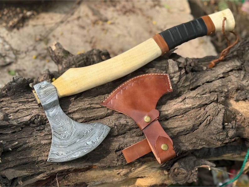 May include: A handcrafted axe with a patterned steel blade and a light-colored wooden handle. The handle is wrapped in black and brown leather. A brown leather sheath with brass hardware protects the blade. The axe rests on a weathered wooden surface.