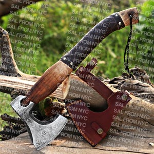 May include: A handcrafted Viking axe with a silver blade featuring intricate engravings. The axe has a wooden handle and a dark brown leather-wrapped grip. Includes a brown leather sheath with a button closure. The axe is designed for display or historical reenactment.