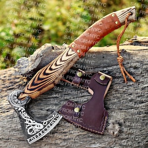 May include: A handcrafted axe and knife set with wooden handles and leather accents. The axe head features intricate engraved designs. The set includes a leather sheath for the axe. The knife has a brown leather-wrapped handle.