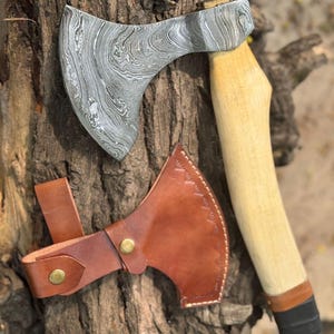 May include: A Damascus steel axe with a wooden handle and a brown leather sheath. The axe head has a unique swirling pattern. The handle is light-colored wood with a black grip. The leather sheath has brass-colored rivets.
