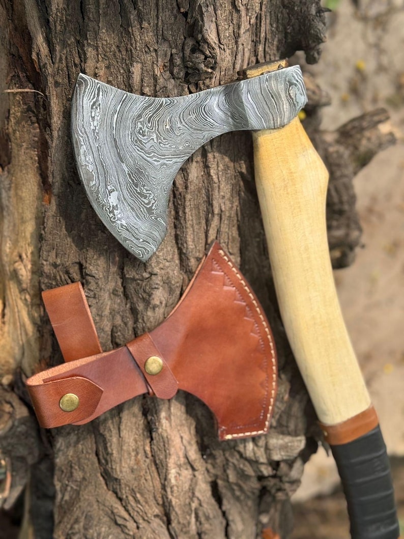 May include: A Damascus steel axe with a wooden handle and a brown leather sheath. The axe head has a unique swirling pattern. The handle is light-colored wood with a black grip. The leather sheath has brass-colored rivets.