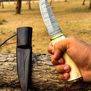 May include: A Damascus steel knife with a cream-colored handle and gold accents is held in a hand. A dark brown leather sheath rests on a log. The knife blade has a unique pattern.