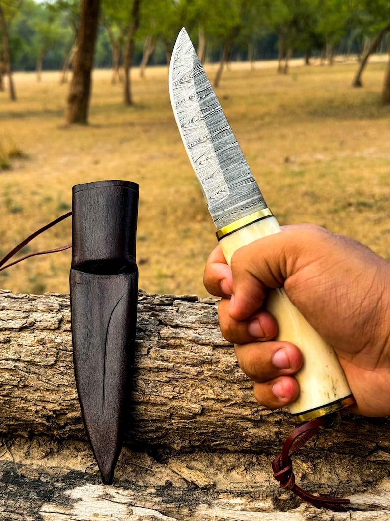 May include: A Damascus steel knife with a cream-colored handle and gold accents is held in a hand. A dark brown leather sheath rests on a log. The knife blade has a unique pattern.