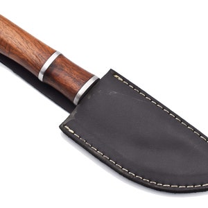 May include: A brown wooden handled knife with a black leather sheath. The sheath has white stitching around the edges.