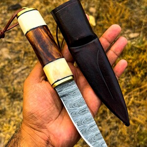 May include: A handcrafted knife with a patterned steel blade and a wooden handle with bone accents. The knife is accompanied by a dark brown leather sheath. The handle features a combination of light and dark wood tones, and the blade has a unique, swirling pattern.