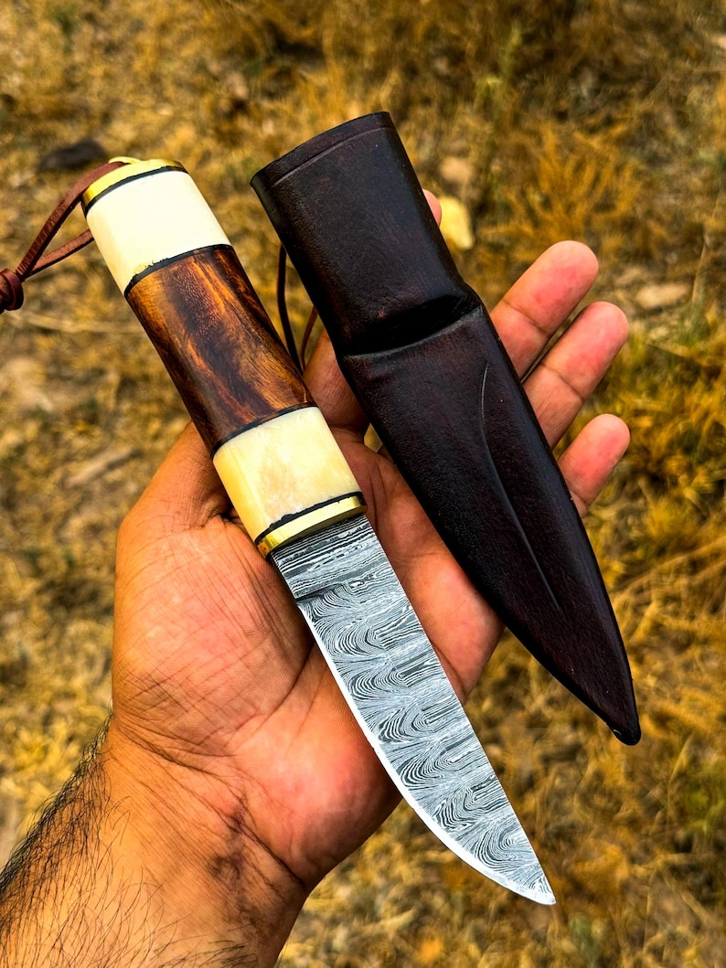 May include: A handcrafted knife with a patterned steel blade and a wooden handle with bone accents. The knife is accompanied by a dark brown leather sheath. The handle features a combination of light and dark wood tones, and the blade has a unique, swirling pattern.
