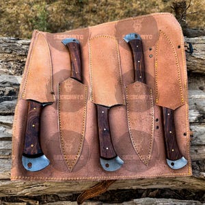 May include: A set of five knives with dark wood handles and silver blades, displayed in a brown leather roll. The handles have a unique grain pattern. The leather roll has the text "SKIEROMYO" and a skull logo.