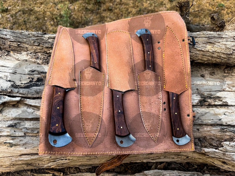 May include: A set of five knives with dark wood handles and silver blades, displayed in a brown leather roll. The handles have a unique grain pattern. The leather roll has the text "SKIEROMYO" and a skull logo.