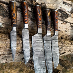 May include: A set of five Damascus steel knives with wooden handles. The blades have a unique swirling pattern, and the handles are a mix of brown, orange, and black. The knives are arranged on a piece of weathered wood.