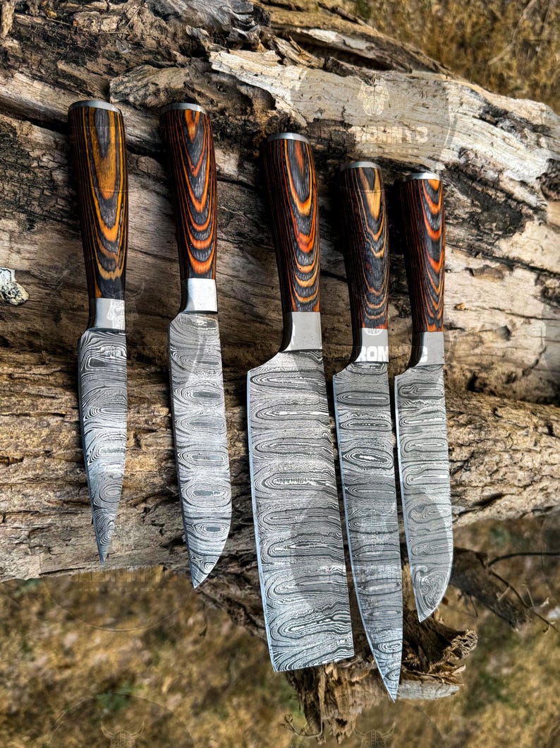 May include: A set of five Damascus steel knives with wooden handles. The blades have a unique swirling pattern, and the handles are a mix of brown, orange, and black. The knives are arranged on a piece of weathered wood.