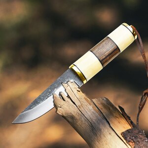 May include: A handmade knife with a wooden and bone handle, featuring a stainless steel blade. The handle has a dark wood centre with cream-coloured sections and gold accents. A leather strap is attached to the end of the handle.