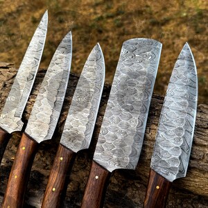 May include: An overhead shot displays five Damascus steel knives with wooden handles. The blades have a unique patterned design. The handles are dark brown with brass rivets. The knives are arranged on a piece of wood.