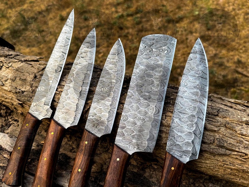 May include: An overhead shot displays five Damascus steel knives with wooden handles. The blades have a unique patterned design. The handles are dark brown with brass rivets. The knives are arranged on a piece of wood.