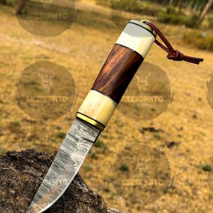 May include: A handcrafted knife with a patterned steel blade and a handle featuring alternating sections of dark wood, bone, and a brass accent. A leather lanyard is attached to the end of the handle. The knife is displayed on a piece of wood.