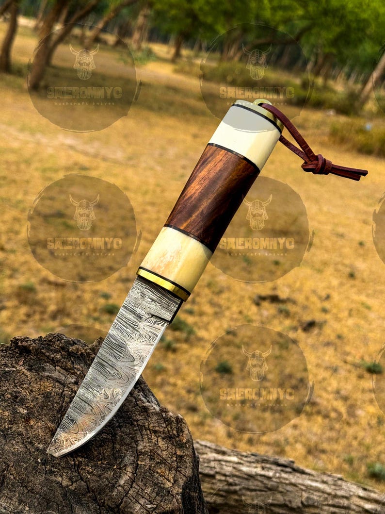 May include: A handcrafted knife with a patterned steel blade and a handle featuring alternating sections of dark wood, bone, and a brass accent. A leather lanyard is attached to the end of the handle. The knife is displayed on a piece of wood.