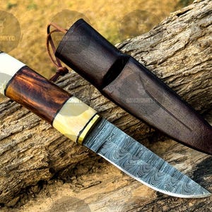 May include: A Damascus steel knife with a wooden handle and leather sheath. The handle features a combination of dark wood, bone, and brass accents. The sheath is dark brown leather. The knife is designed for outdoor use.