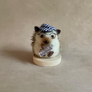 May include: A handmade hedgehog ornament wearing a blue and white checked cap and round spectacles. The hedgehog holds a miniature newspaper, "The New York Times", and sits on a small wooden base. The figurine has a neutral colour palette with brown and beige tones.