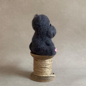 May include: A dark grey felt hippo figure with a pink mouth and small black eyes. The hippo sits on a wooden spool wrapped with natural twine. The background is a plain, neutral colour.