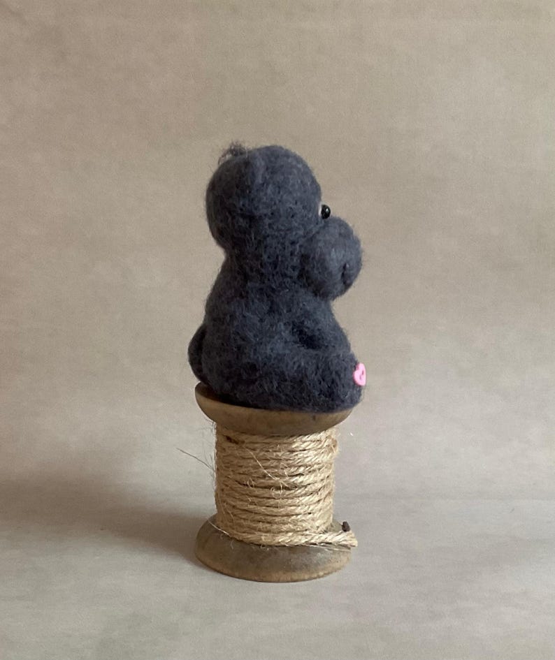 May include: A dark grey felt hippo figure with a pink mouth and small black eyes. The hippo sits on a wooden spool wrapped with natural twine. The background is a plain, neutral colour.