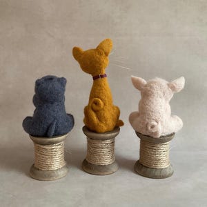 May include: Three felt animal figures: a blue hippo, a yellow cat with a purple collar, and a white pig. Each sits on a wooden spool. The hippo and pig face away, while the cat looks to the side.