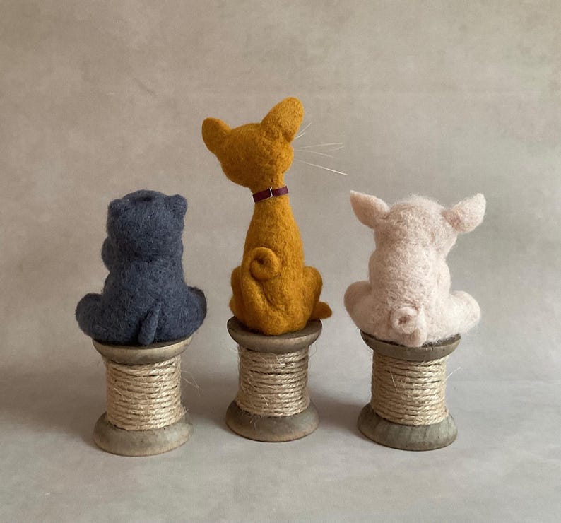 May include: Three felt animal figures: a blue hippo, a yellow cat with a purple collar, and a white pig. Each sits on a wooden spool. The hippo and pig face away, while the cat looks to the side.
