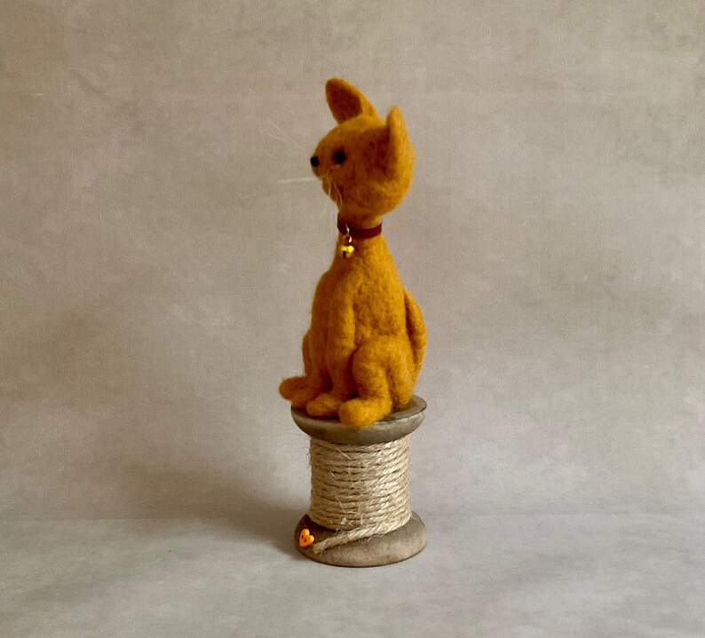 May include: A handcrafted orange cat figure with a red collar and a small gold bell, perched on a wooden spool wrapped with twine. The cat has pointed ears and white whiskers. A unique decorative item.