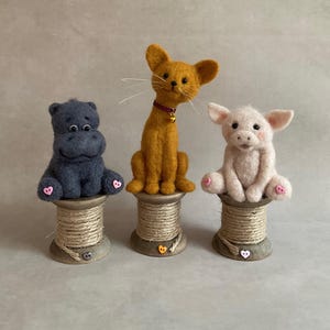 May include: Three handmade felt animal figurines: a grey hippo, an orange cat with a red collar, and a pink pig. Each sits atop a wooden spool. The hippo and pig have pink accents on their feet.