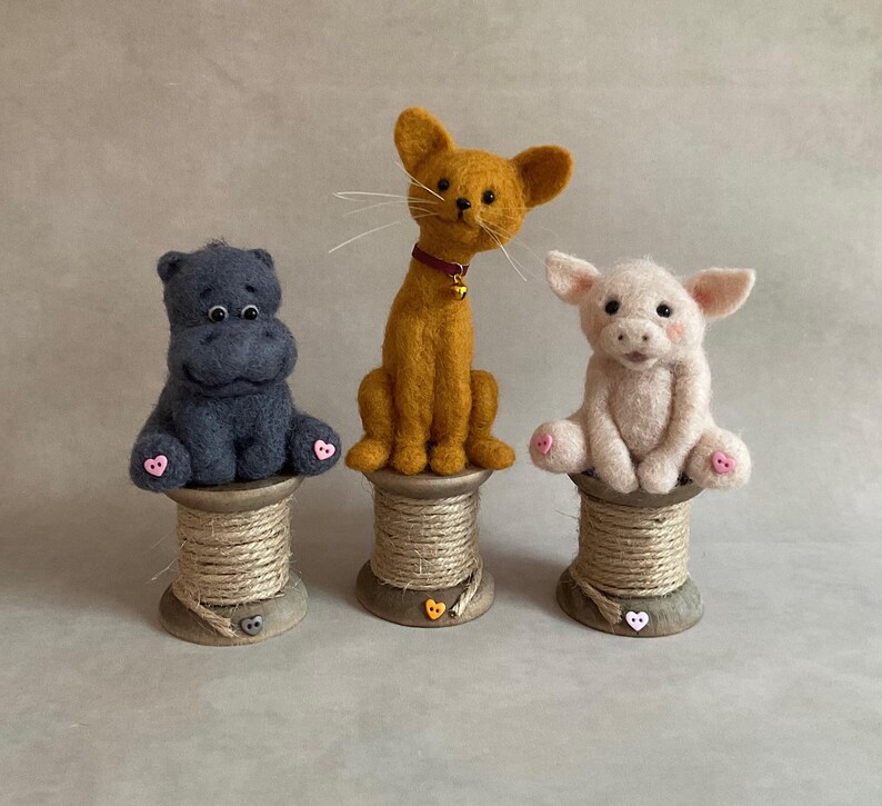 May include: Three handmade felt animal figurines: a grey hippo, an orange cat with a red collar, and a pink pig. Each sits atop a wooden spool. The hippo and pig have pink accents on their feet.