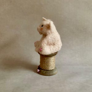 May include: A light pink felt pig figurine with a small horn, sitting on a wooden spool wrapped in twine. The pig has pink accents on its ears and feet. A small pink heart is attached to the base of the spool.