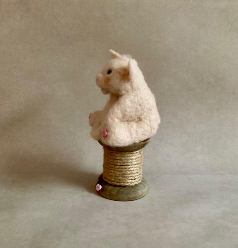 May include: A light pink felt pig figurine with a small horn, sitting on a wooden spool wrapped in twine. The pig has pink accents on its ears and feet. A small pink heart is attached to the base of the spool.
