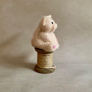 May include: A light pink, felted pig figurine with black eyes and pink accents on its ears and feet. The pig is seated on a wooden spool wrapped with natural twine. The background is a neutral, solid colour.
