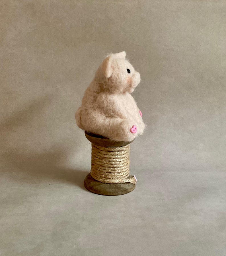 May include: A light pink, felted pig figurine with black eyes and pink accents on its ears and feet. The pig is seated on a wooden spool wrapped with natural twine. The background is a neutral, solid colour.