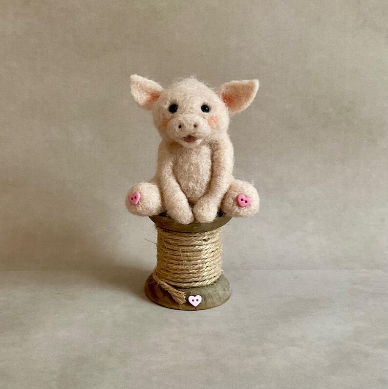 May include: A light pink felt piglet with black eyes, sitting on a wooden spool wrapped in twine. The piglet has pink accents on its ears, cheeks, and feet. A small, pink heart-shaped button is on the spool.