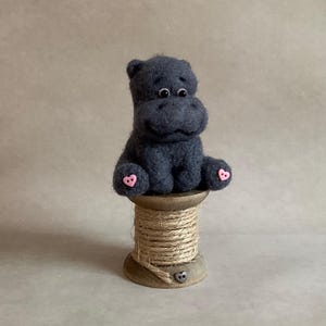 May include: A handcrafted, dark grey felt hippo figure with pink heart-shaped details on its feet. The hippo is perched on a wooden spool wound with natural twine. The background is a plain, neutral colour.
