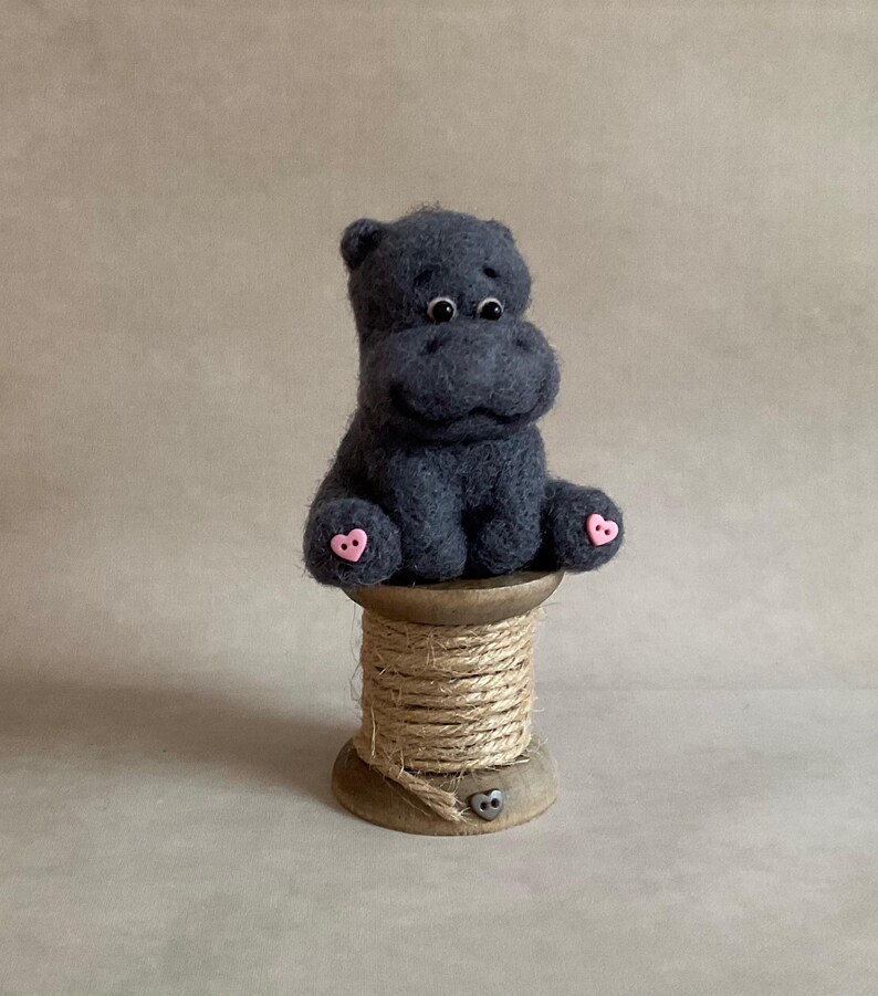 May include: A handcrafted, dark grey felt hippo figure with pink heart-shaped details on its feet. The hippo is perched on a wooden spool wound with natural twine. The background is a plain, neutral colour.