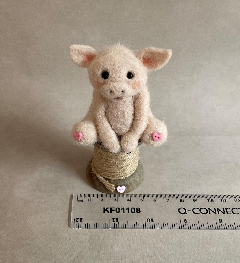 May include: A light pink felt piglet figurine with black eyes and pink accents. The piglet sits on a twine-wrapped spool, with a heart-shaped button. A ruler with the text "KF01108" is in the foreground.